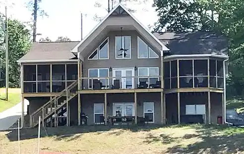 Two-story house with a large covered porch, multiple windows, screened sections, and outdoor seating, surrounded by grass and trees.