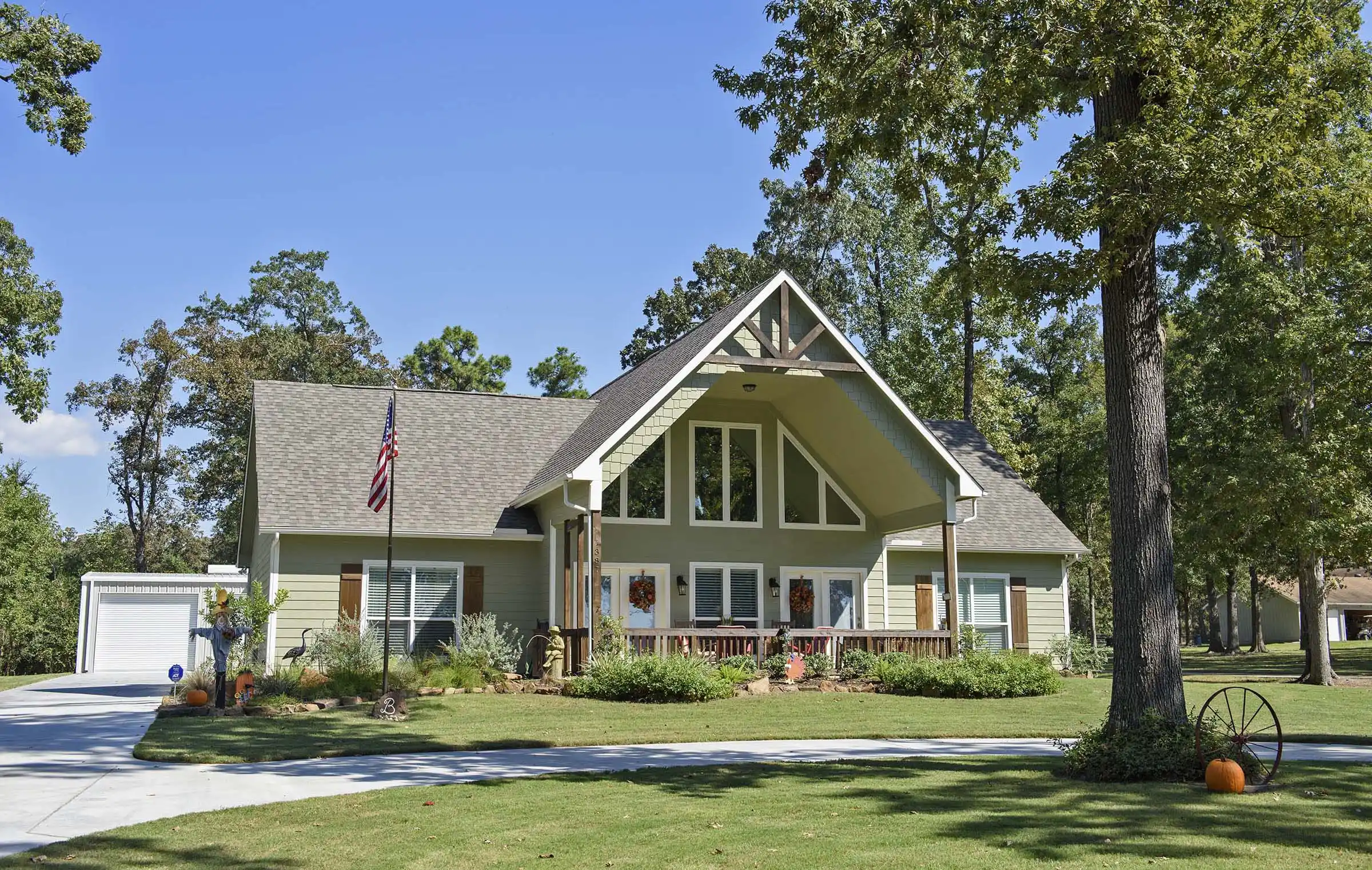 Single-story house with a front porch, American flag, large tree, pumpkins, and a well-kept lawn under a clear sky.