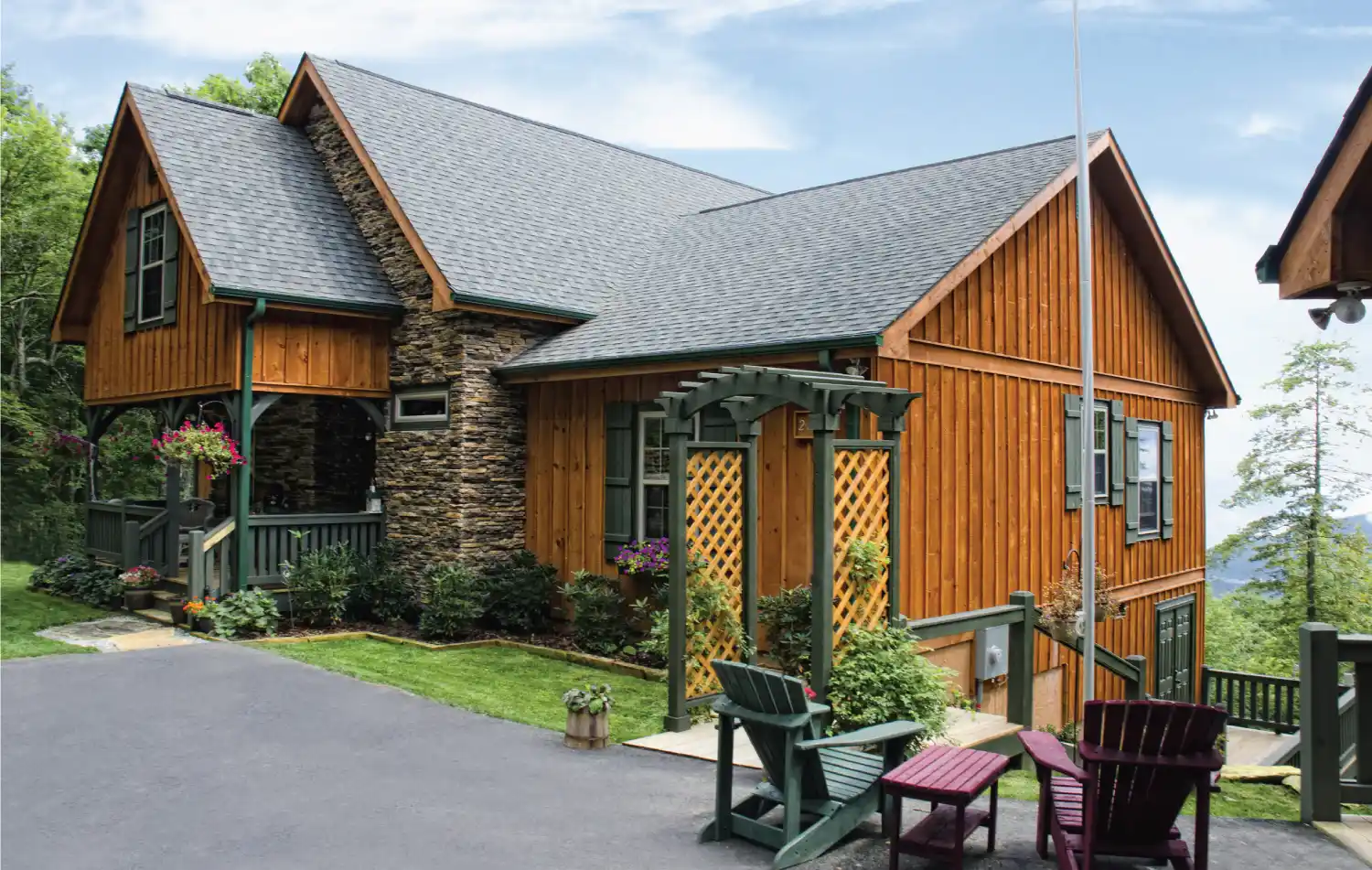 Wood-sided house with stone accents, a covered porch, pergola, green and red outdoor chairs, potted plants, and a paved driveway surrounded by trees.