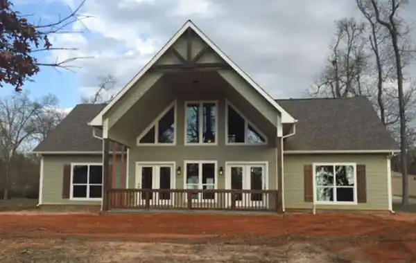 Front view of a house with a large covered porch, multiple windows with white frames, and a gabled roof, surrounded by bare trees and dirt ground.