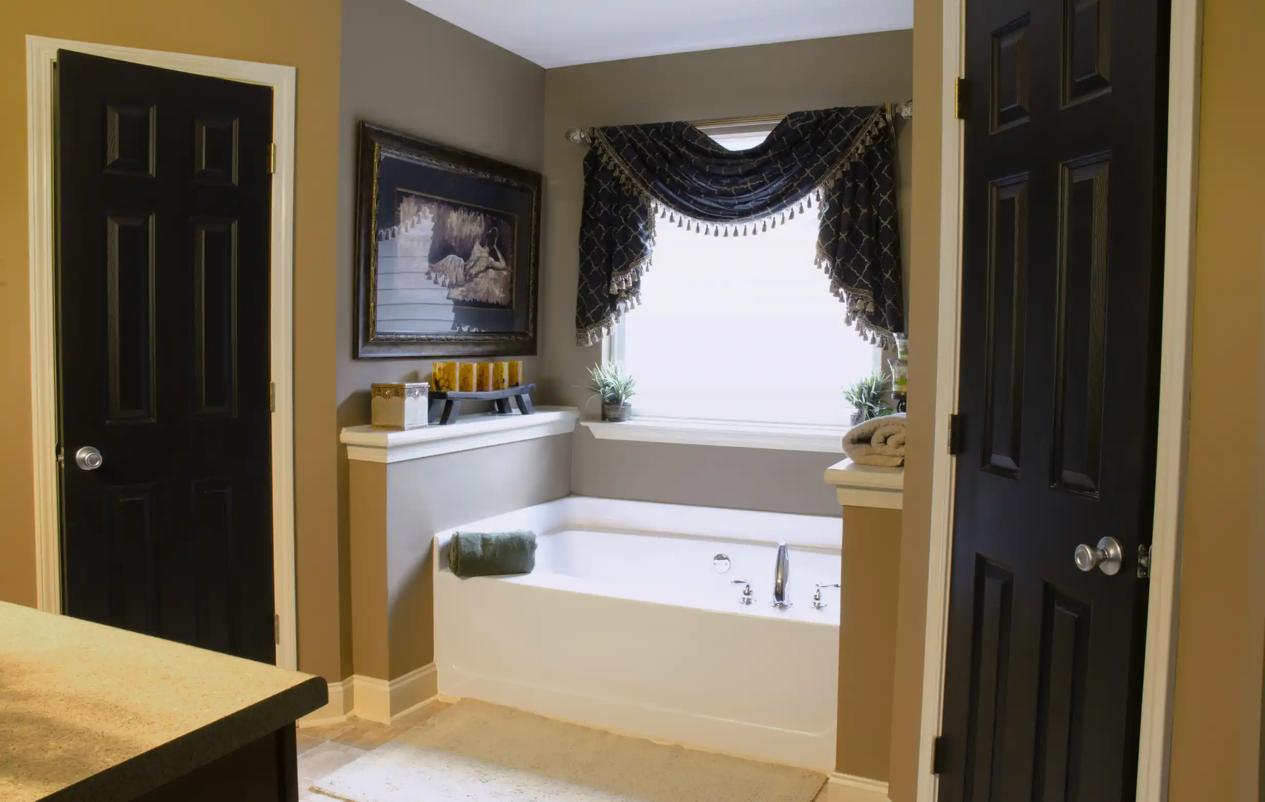 Bathroom with a white bathtub under a window with dark curtains, two potted plants, a towel on the tub edge, and dark doors on either side.