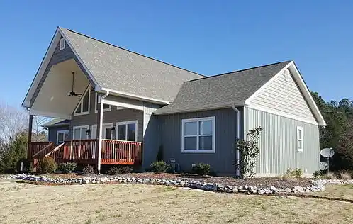 Gray house with white trim, covered front porch, wooden deck, several windows, and a landscaped yard with rocks under a clear blue sky.