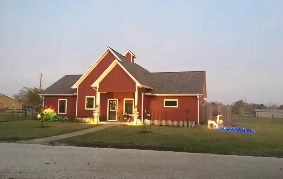 Red house with white trim, a front porch, small white fence, lawn, and inflatable decorations, with trees and a cloudy sky in the background.