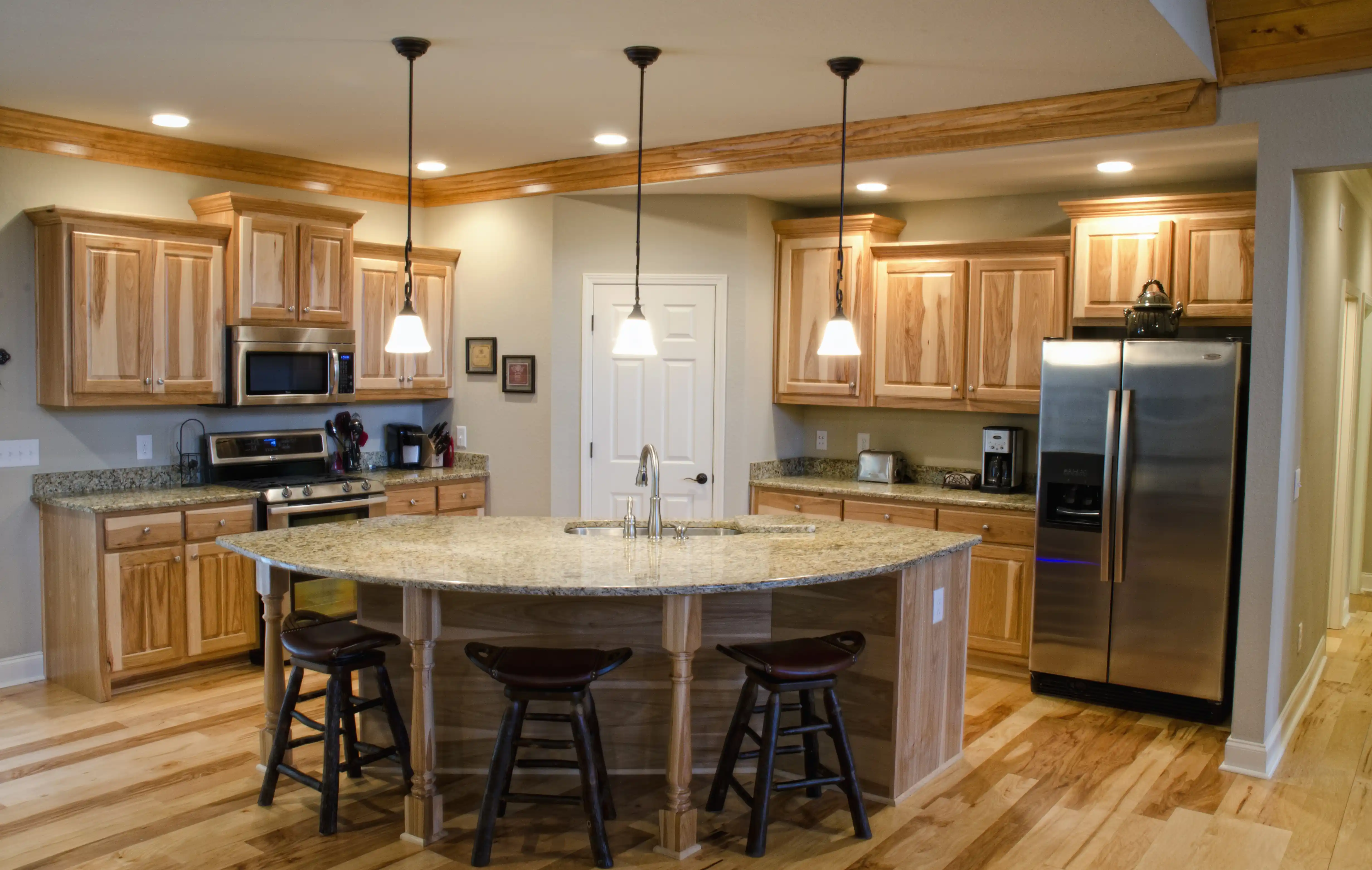 Spacious kitchen with a large island featuring a stone countertop, built-in sink, three stools, stainless steel appliances, and wooden cabinets.
