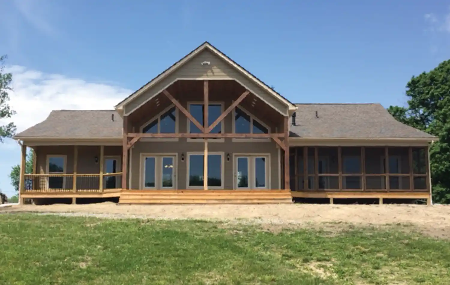 Single-story house with a large covered porch, multiple windows and doors, wood siding, and a grassy lawn under a clear blue sky.