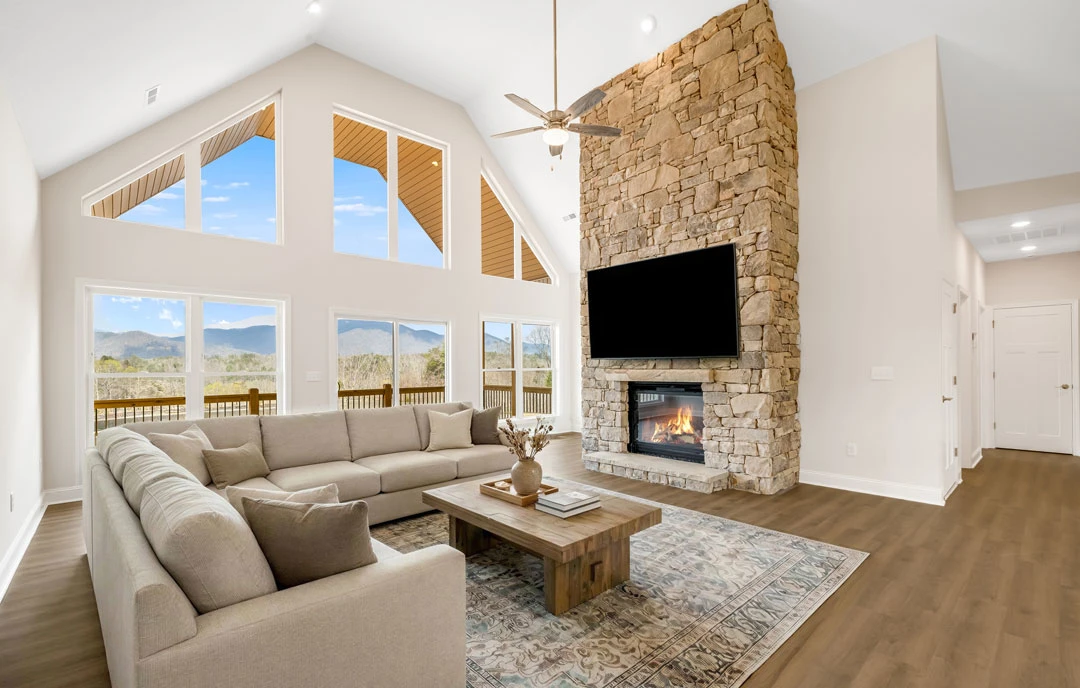Living room with a stone fireplace, a black TV mounted above, a gray couch with pillows, a coffee table with a vase, and a white door with a gold handle.