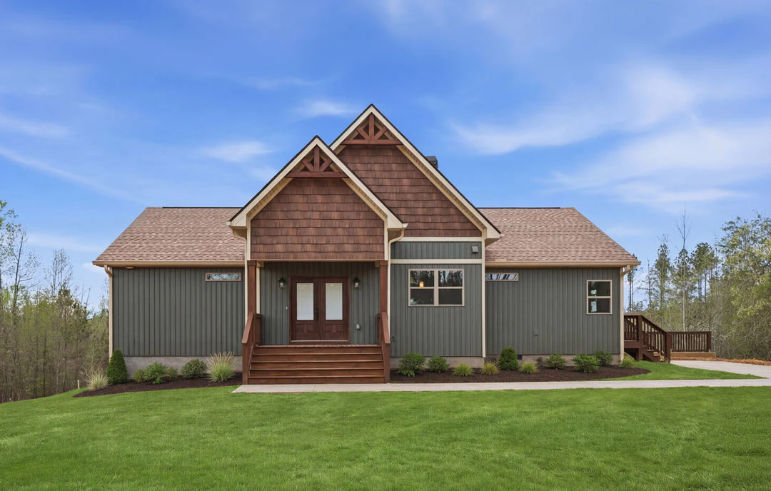 Two-story farmhouse with light siding, double glass front doors, a covered porch, brown wooden staircase with railing, and a grass lawn bordered by a wooden fence under a blue sky.