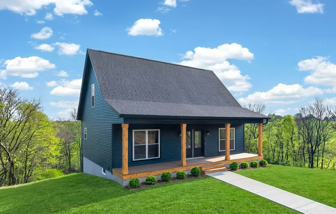 Blue house with white-framed windows, a covered porch, and a grassy lawn bordered by mature trees; a walkway leads from the yard to the entrance.