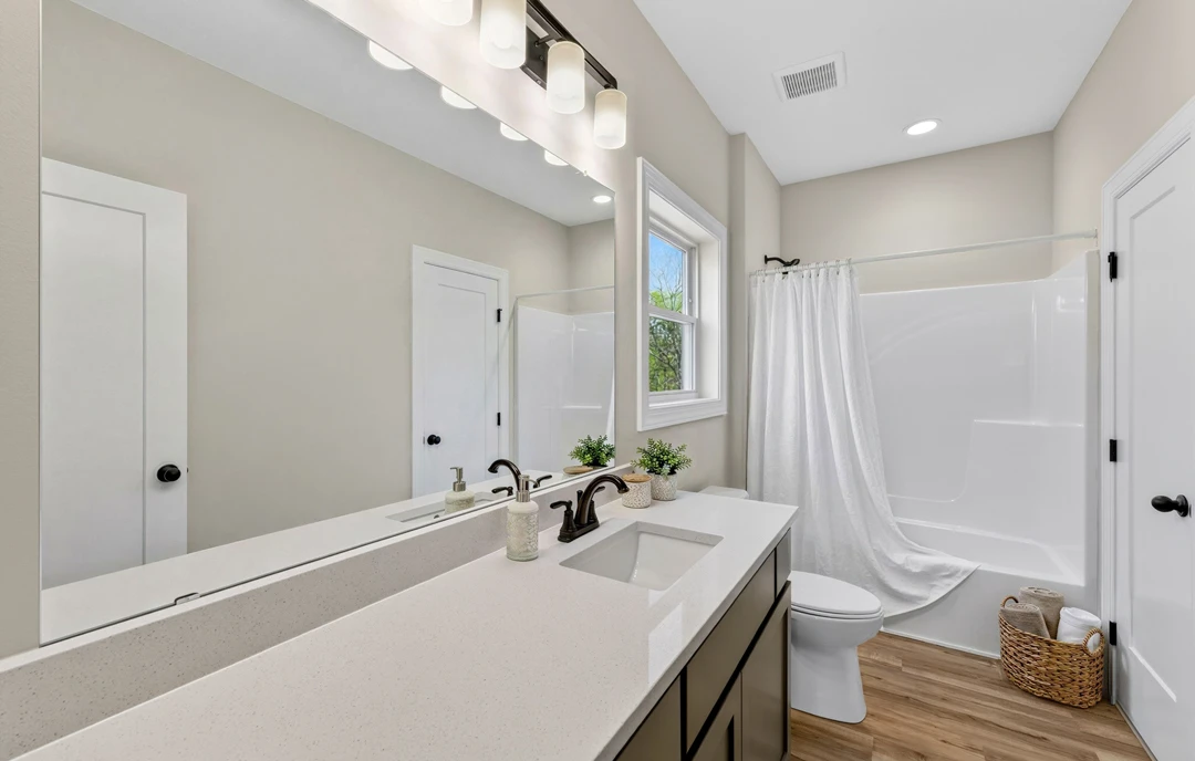 Bathroom with a white shower curtain, white sink, and basket of rolled towels on tile flooring; toilet and cabinetry are visible near a wall-mounted mirror.