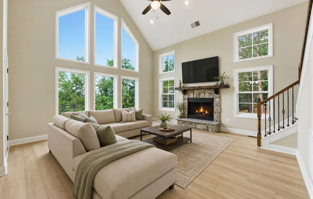 Living room with hardwood flooring, a fireplace with fire, large windows, a gray couch, a coffee table with a tray, and a staircase visible in the background.