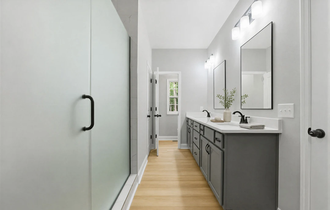 Bathroom with a white sink and countertop set in wood cabinetry, large mirror above, wood flooring, a white door with black handle, and a small plant in a vase on the counter.