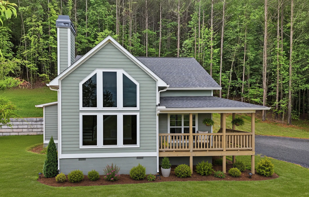 White farmhouse-style home with a covered front porch, multiple windows with white frames, and a landscaped yard bordered by trees and grass.