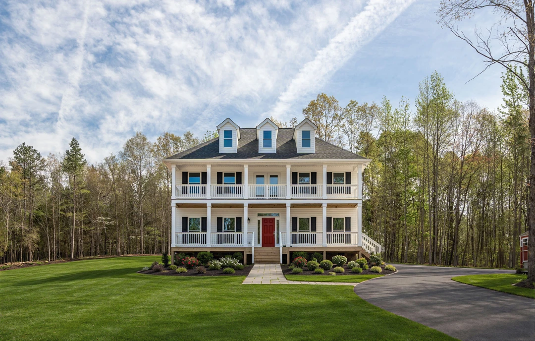 Large white house with a red front door, multiple dormer windows on the roof, a driveway and walkway leading to the entrance, and a lawn bordered by trees and shrubs.