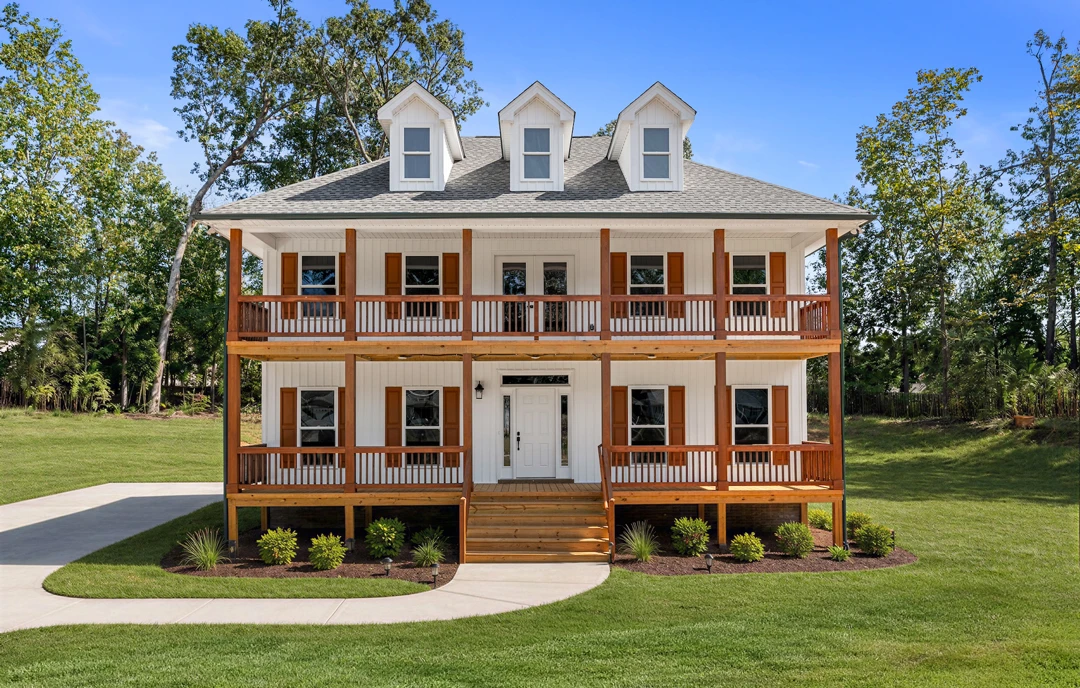 White house with brown railings, a covered porch with potted plants, wooden deck and staircase, surrounded by a green lawn and mature trees.