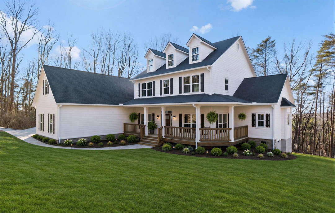Large white house with a covered porch, wooden railing, and expansive lawn, surrounded by mature trees under a blue sky.