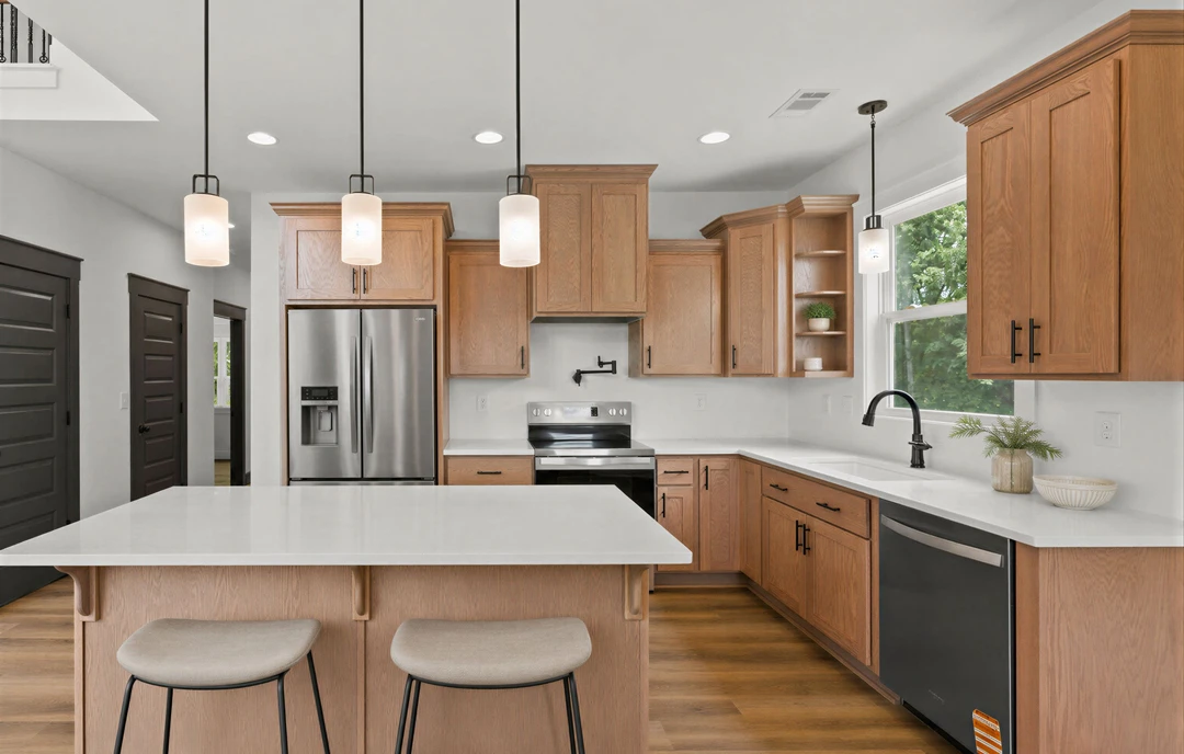 Kitchen with wood cabinets, white granite countertop, black dishwasher with silver handle, stainless refrigerator, two cushioned chairs at the counter, and a plant in a vase.