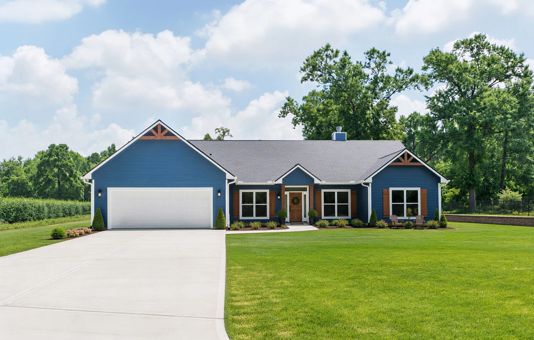 Blue house with white-framed windows, a front door, attached garage, and a driveway bordered by grass and flowers; lawn with outdoor chairs visible in the background.