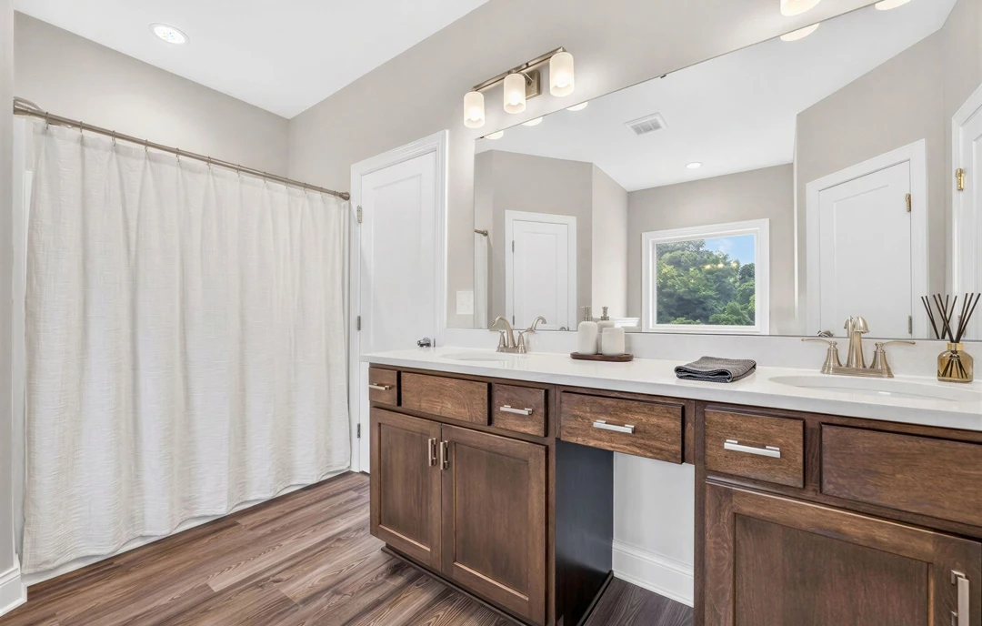 Bathroom with a large wall mirror above a white sink and cabinet, white tile flooring, a white curtain on a rod, and a window showing trees outside.