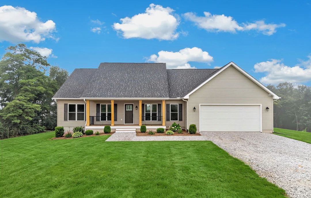 White house with a covered porch, white garage door, stone path across a green lawn, and a gravel driveway bordered by mature trees.