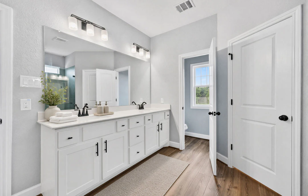 Bathroom with white cabinets and white countertops, a large mirror above the sink, white tile walls, a plant in a white vase, and a window showing trees outside.