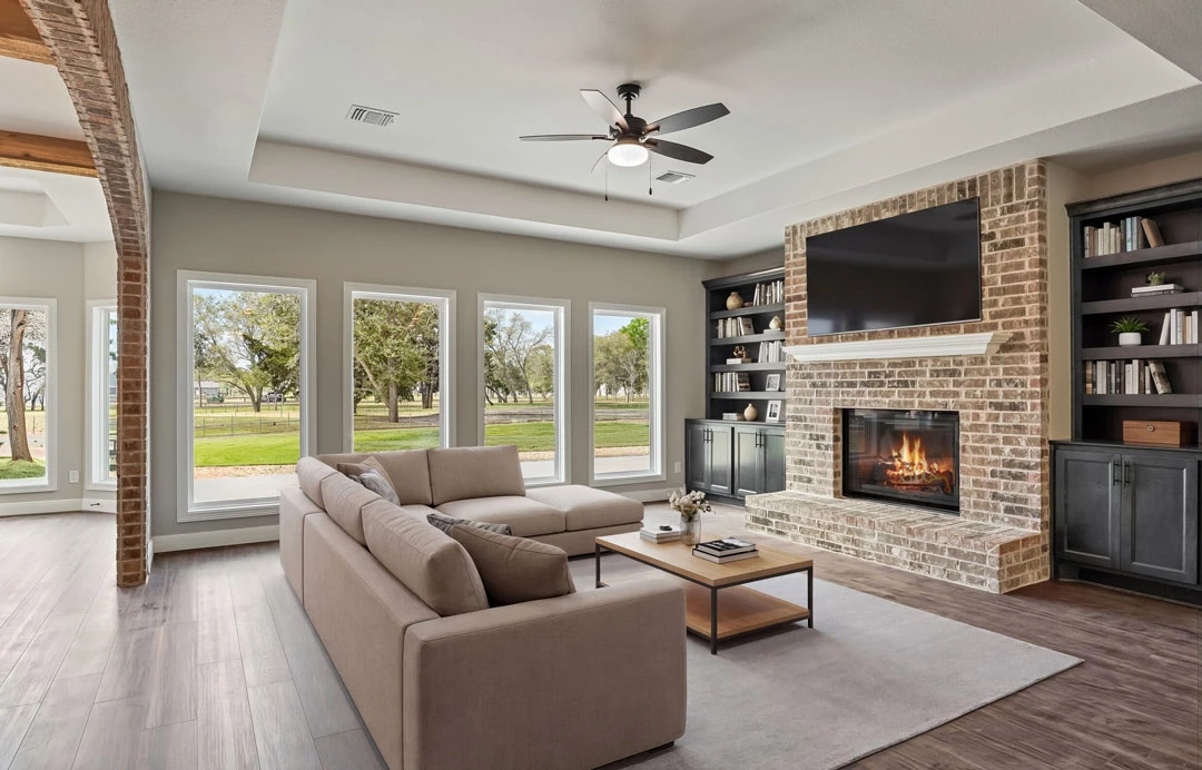 Living room with a wood-burning fireplace, light-colored couch, wood flooring, coffee table with books, ceiling fan with light, and a wall-mounted mirror.