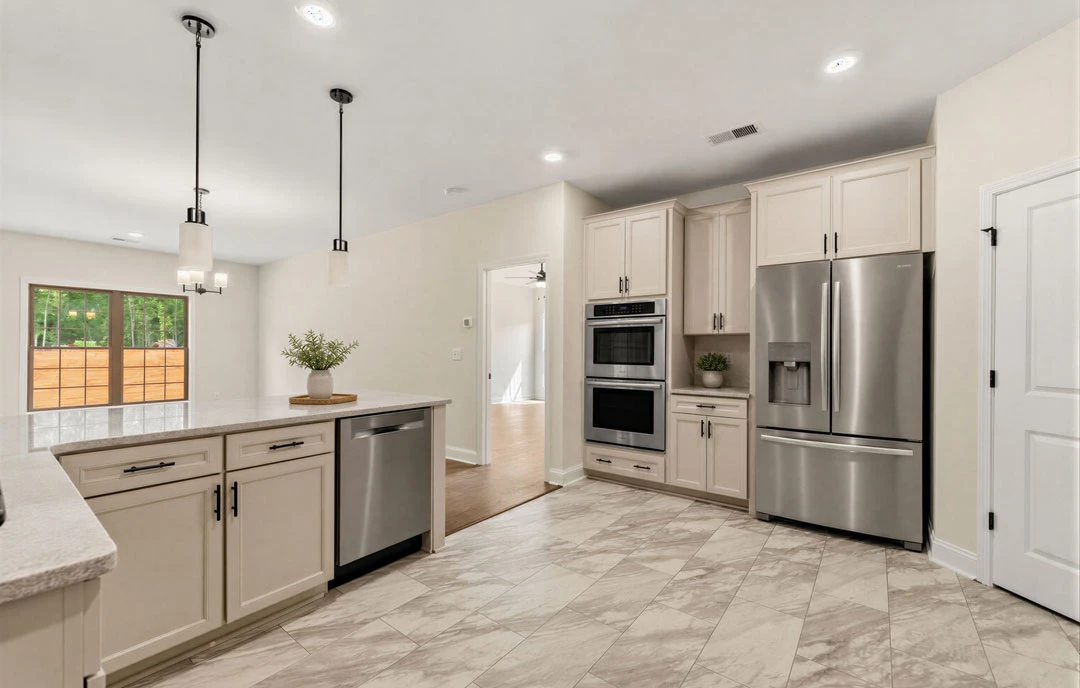Kitchen with stainless steel appliances, including a refrigerator and oven, white cabinetry, granite countertops, wood flooring, a plant in a vase, and a window overlooking trees.
