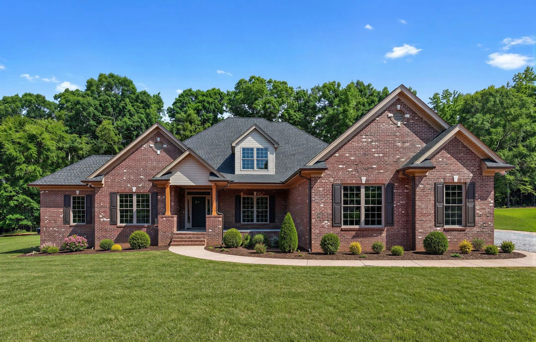 Red brick house with multi-pane windows, surrounded by a green lawn and mature trees in the background.