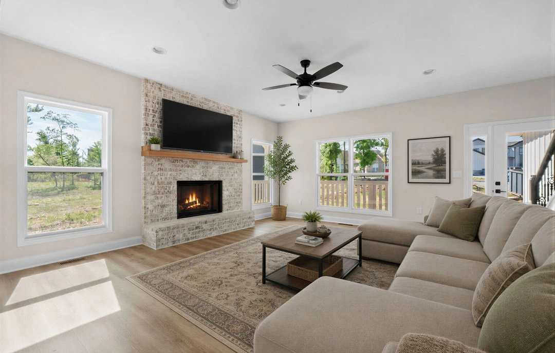 Living room with a brick fireplace, a couch, a black television mounted above the hearth, ceiling fan with light, coffee table, and pillows; framed landscape art and a window are