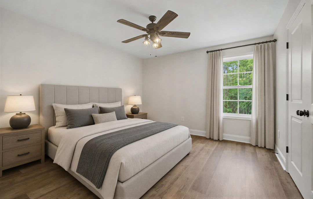 Bedroom with a ceiling fan, a bed featuring a grey blanket and pillows, a window with curtains showing trees outside, a lamp with a white shade, and a bowl atop a dresser.