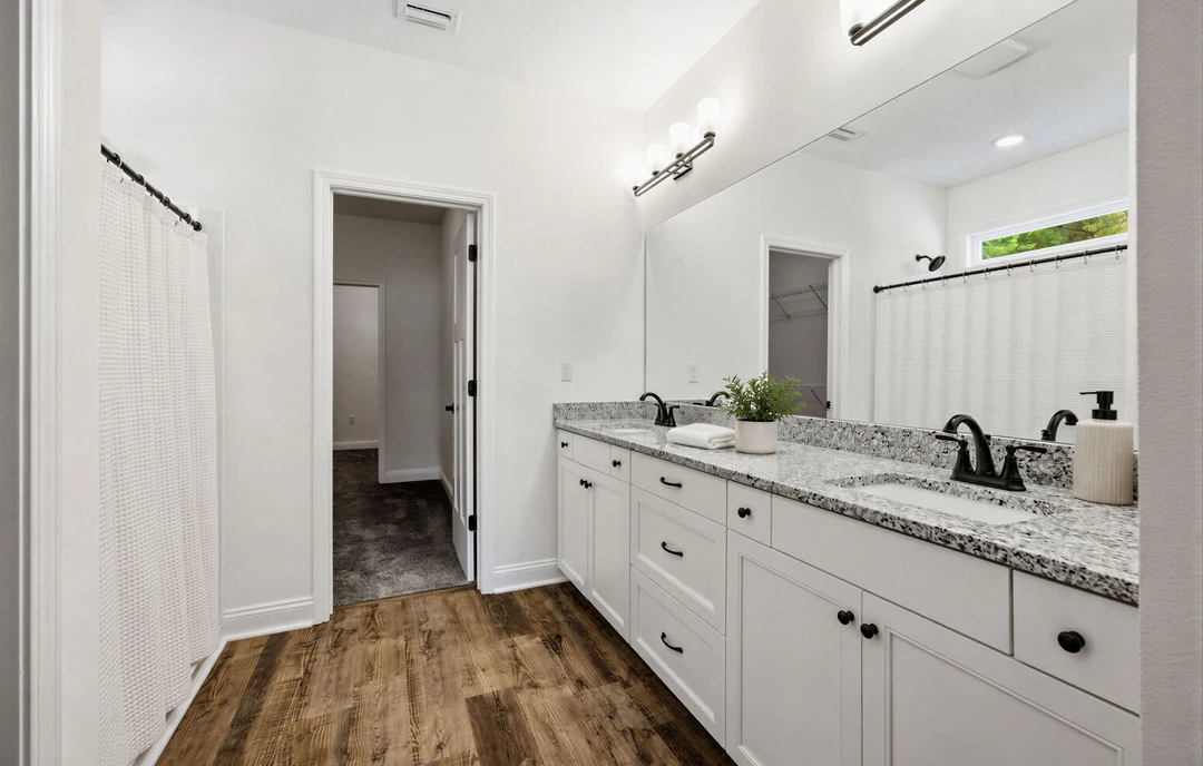 Bathroom with a large wall mirror, white cabinets, wood flooring, a potted plant on the countertop, white soap dispenser with black pump, and a white towel near a doorway.