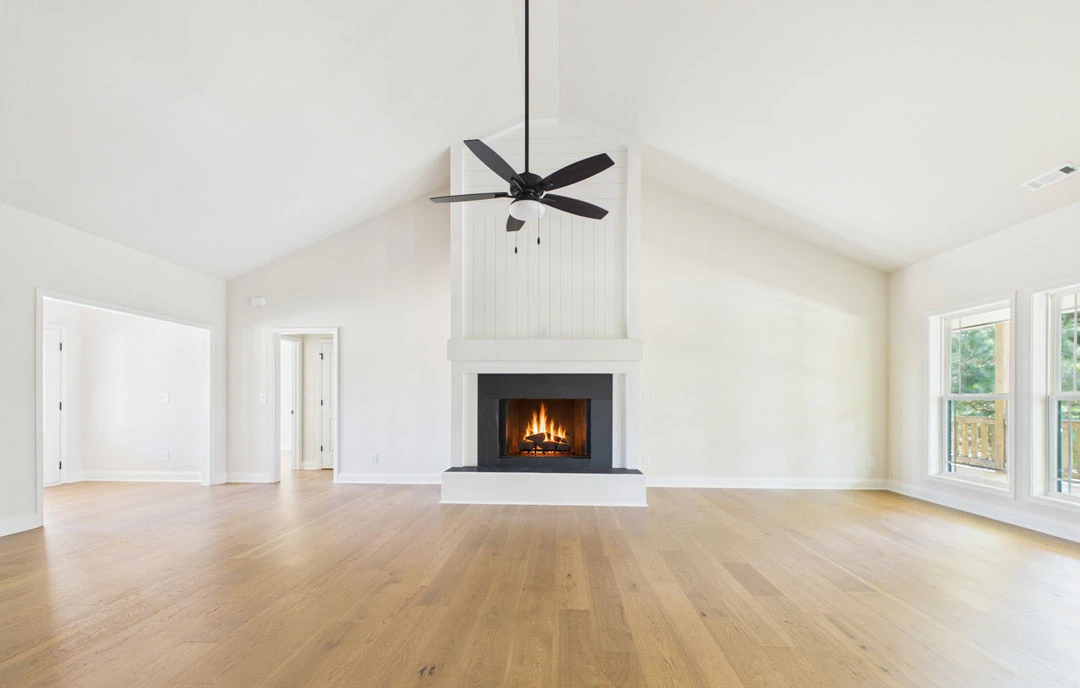 Living room with hardwood floors, a fireplace with a burning fire, a ceiling fan with a light fixture, and a window overlooking the outdoors.