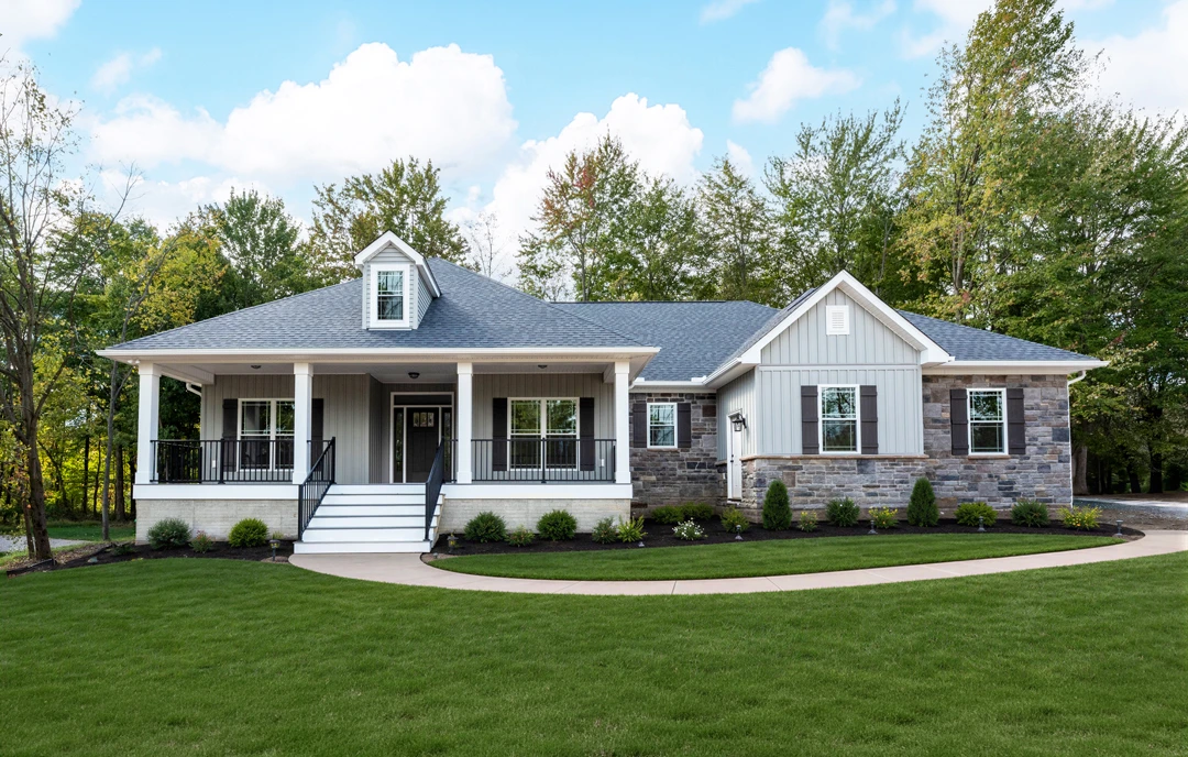 Two-story farmhouse with white siding, a covered front porch featuring a stone wall, black door with white trim, and a lawn with a concrete walkway bordered by mature trees.