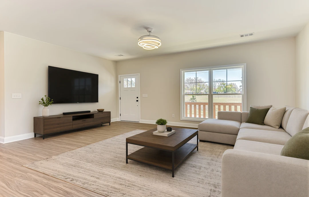 Living room with a brown wooden entertainment center, black TV screen, gray couch with cushions, coffee table holding a plant, laminate wood flooring, and a window showing a field