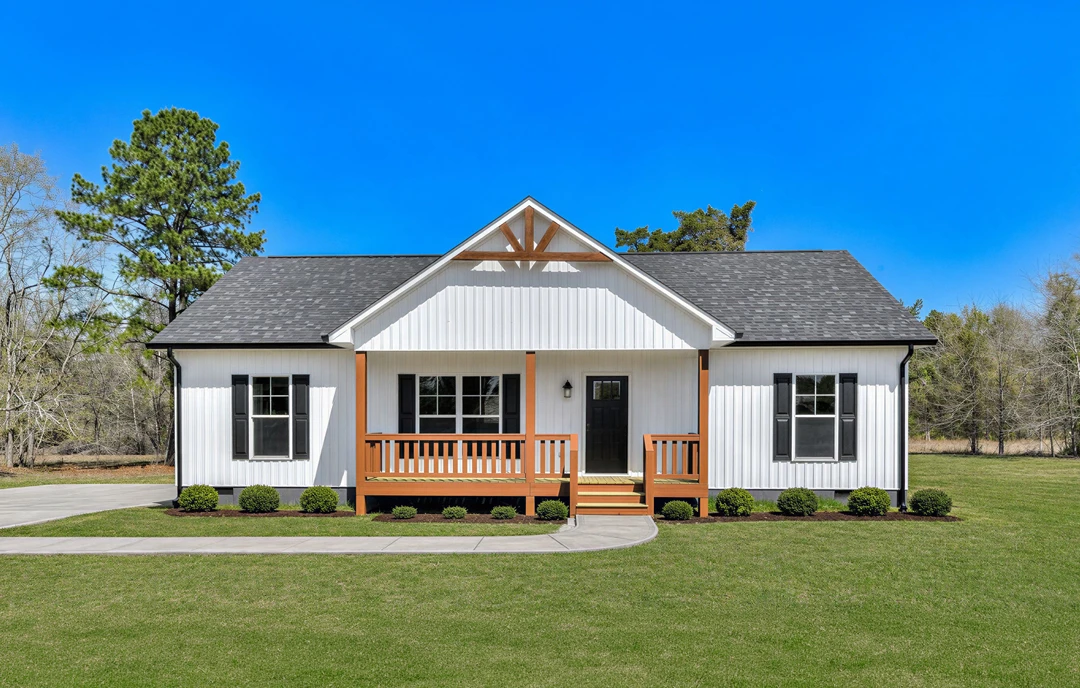 White farmhouse-style exterior with black front door, wooden porch railing, and a well-kept lawn; a tree and green plants are visible near the wooden deck.