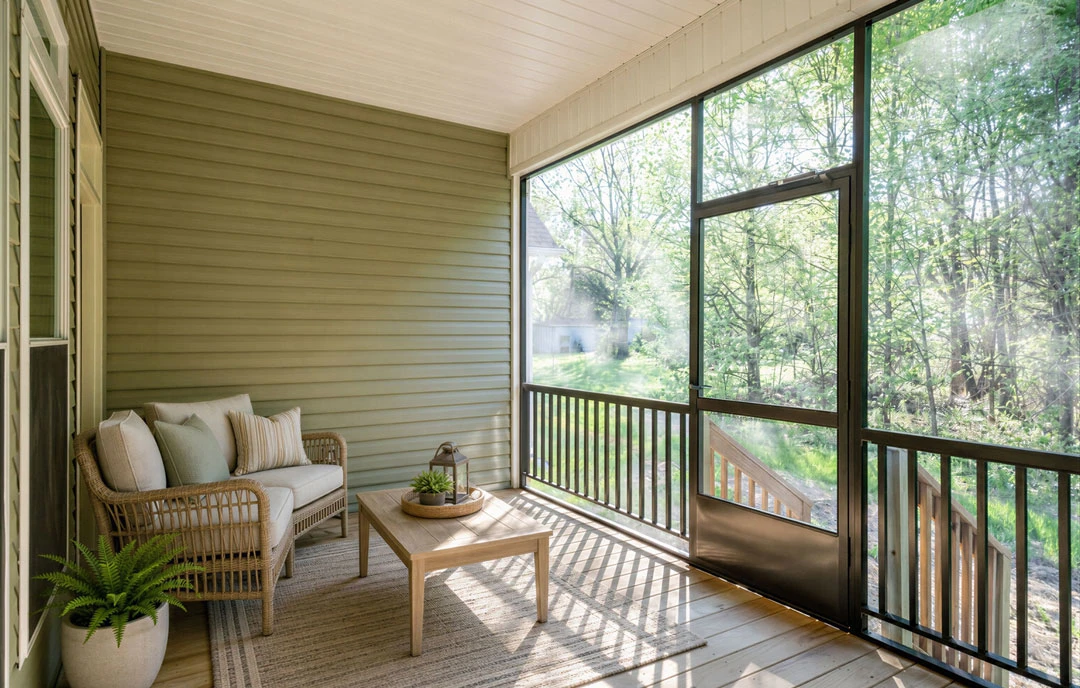 Covered porch with a light-colored couch, coffee table, potted fern, and large glass door leading inside.