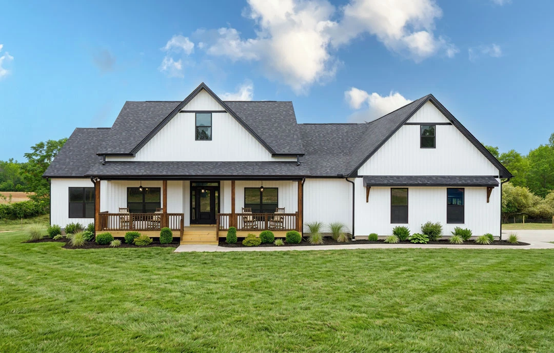 White farmhouse-style exterior with horizontal siding, covered front porch, wooden deck with railing, and a lawn with a few bushes under a blue sky with scattered clouds.