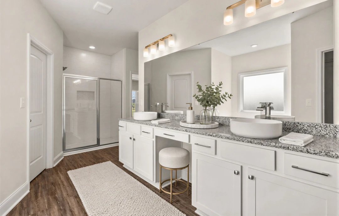 Bathroom with a wood vanity, white countertop and sink, large mirror above, glass vase with a plant, glass shower enclosure, white rug on wood flooring, and a cushioned stool.