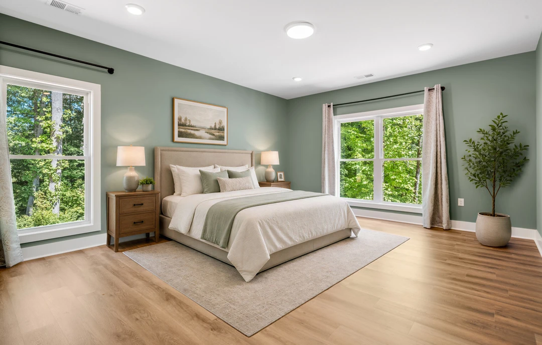 Bedroom with a bed featuring a white bedspread, a window with coverings, wood flooring, a nightstand with a white lamp, a dresser, a potted tree, and a painting on the wall.