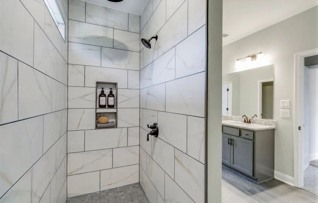 Bathroom with a tiled shower wall, shower head, granite countertop sink, and brown bottles on a shelf; white door frame and border visible.