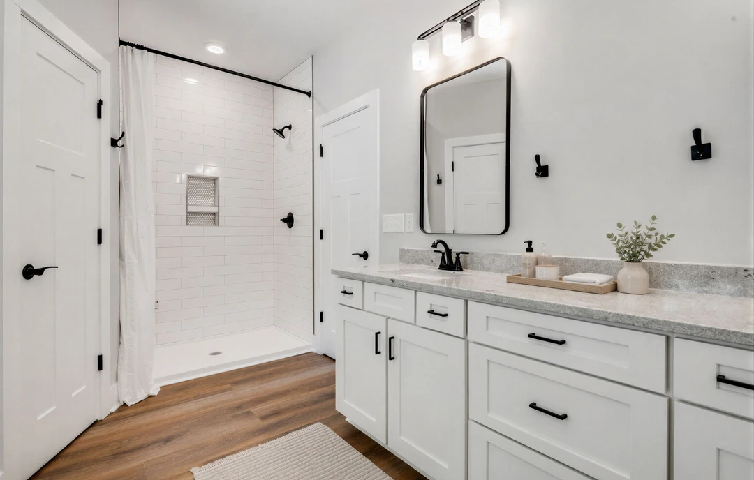 Bathroom with a glass-enclosed shower, large mirror above a white vanity with black hardware, wood flooring, and a potted plant in a vase on the countertop.