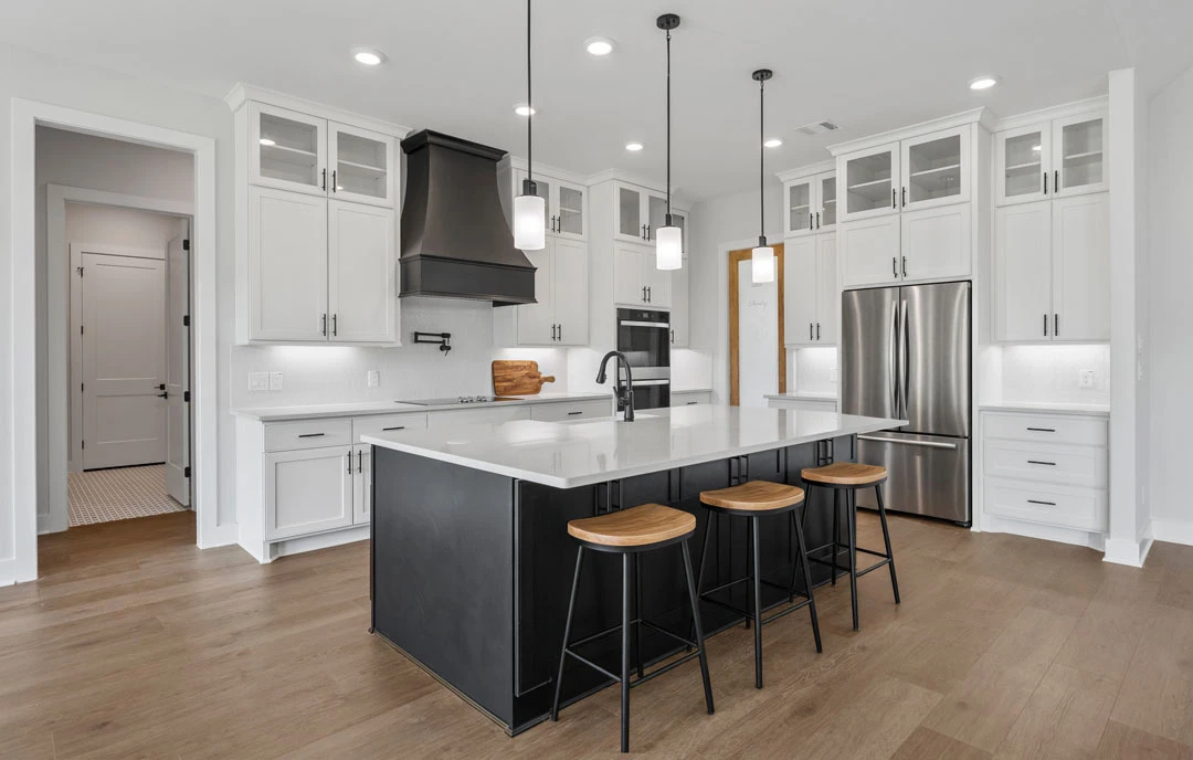 Kitchen with a large central island featuring wood seat bar stools, black hood above the cooktop, stainless steel refrigerator with black handles, and granite countertops on