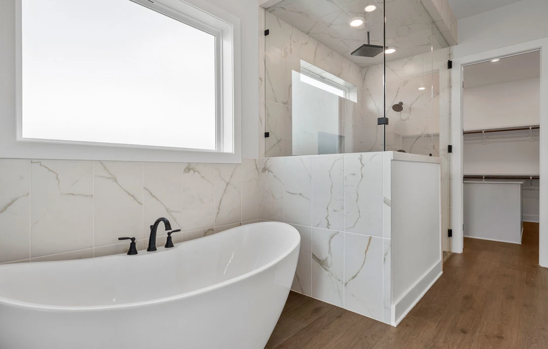 Bathroom with a white freestanding bathtub, glass-enclosed shower, black faucet on a white stone countertop, and a white shelf with a wooden surface beneath a window.