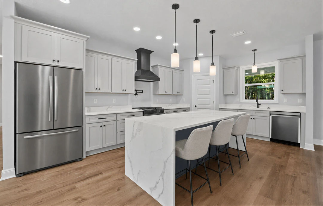 Kitchen with white cabinets, a granite island countertop with chairs, stainless steel refrigerator, black stove hood, and a window with a hanging light fixture.