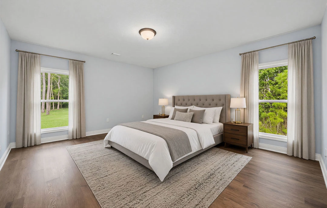 Bedroom with a large bed featuring a headboard, white bedding, and pillows, a nightstand with a lamp, a rug on the floor, and windows overlooking trees and grass.