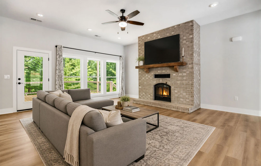 Living room with a gray couch draped with a blanket, a brick fireplace with a fire burning, wood or laminate flooring, a ceiling fan with a light, and a television mounted above