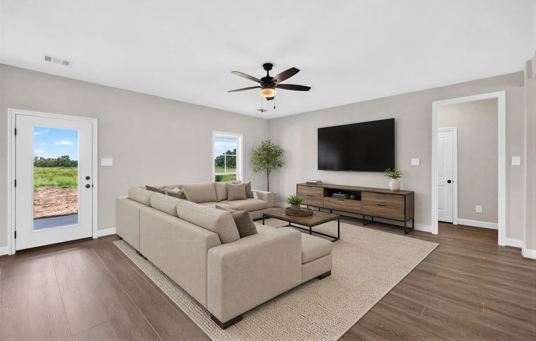 Living room with a gray couch, wall-mounted television, ceiling fan with light, wood laminate flooring, and a potted tree near a door overlooking a field and trees.