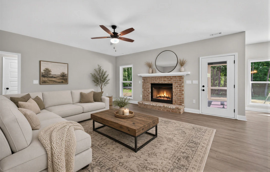 Living room with wood flooring, a gray couch with pillows, a coffee table holding a plant and candle, a fireplace with a hearth, a ceiling fan with a light, a painting of trees on