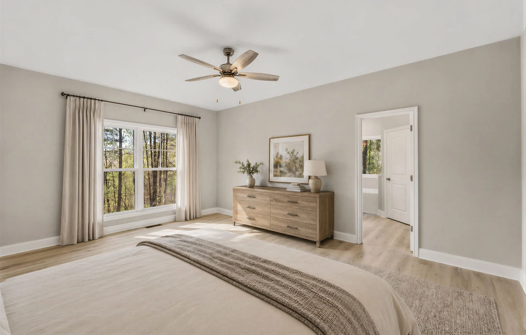 Bedroom with a large bed, white linens, a wooden dresser topped with a white vase, ceiling fan with light, nightstand with a white-shaded lamp, and a window showing trees outside.