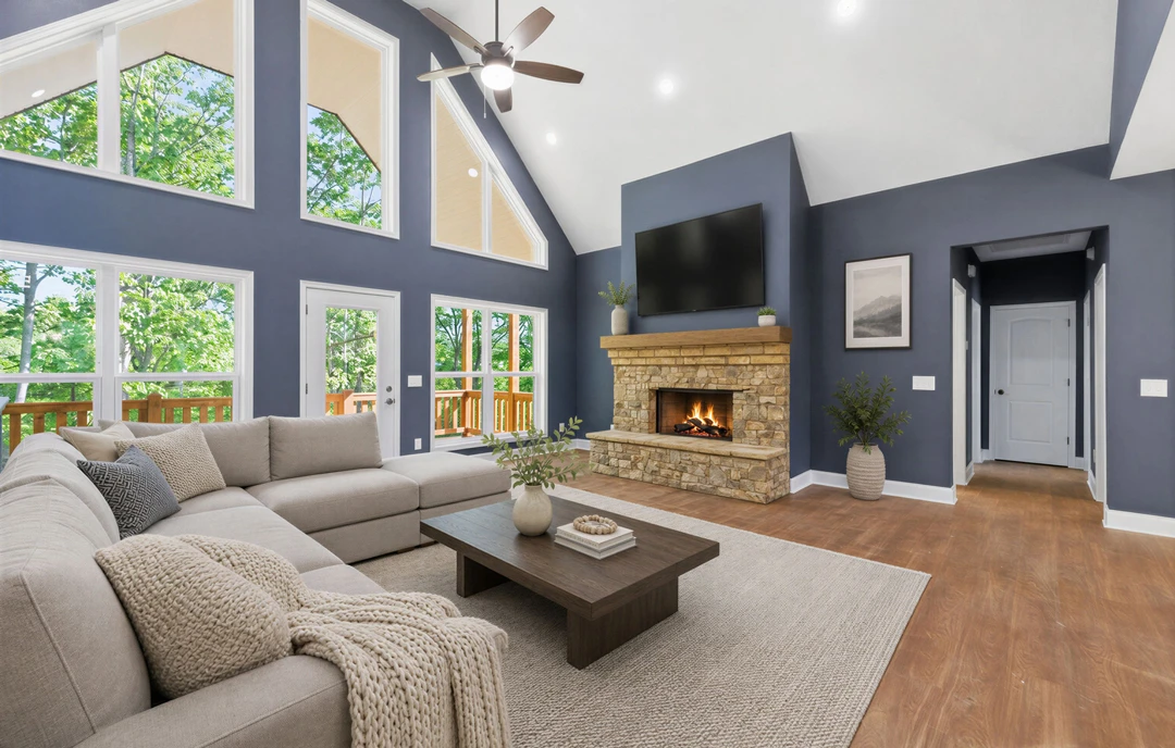 Living room with wood flooring, a fireplace with a fire, a ceiling fan, a couch with pillows and a blanket, a coffee table holding books and a plant, and additional potted plants.