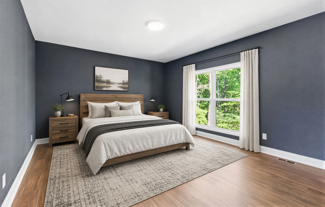 Bedroom with a wood headboard bed, pillows, and a nightstand with a lamp, featuring laminate or hardwood flooring, a dresser, a river painting, and a window with trees visible